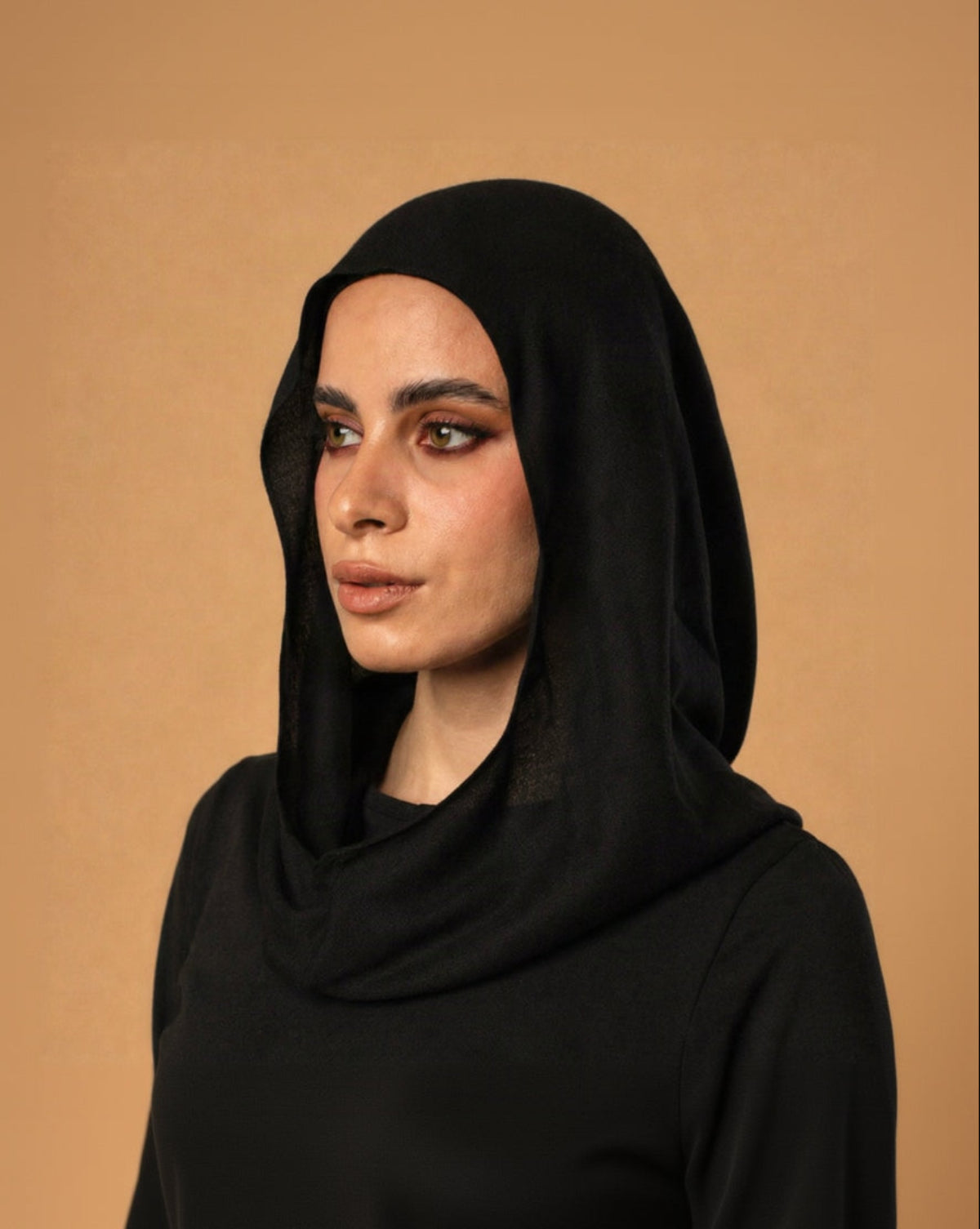 A woman wearing a black draped headscarf or hijab, shown from the chest up against a plain warm beige studio background.